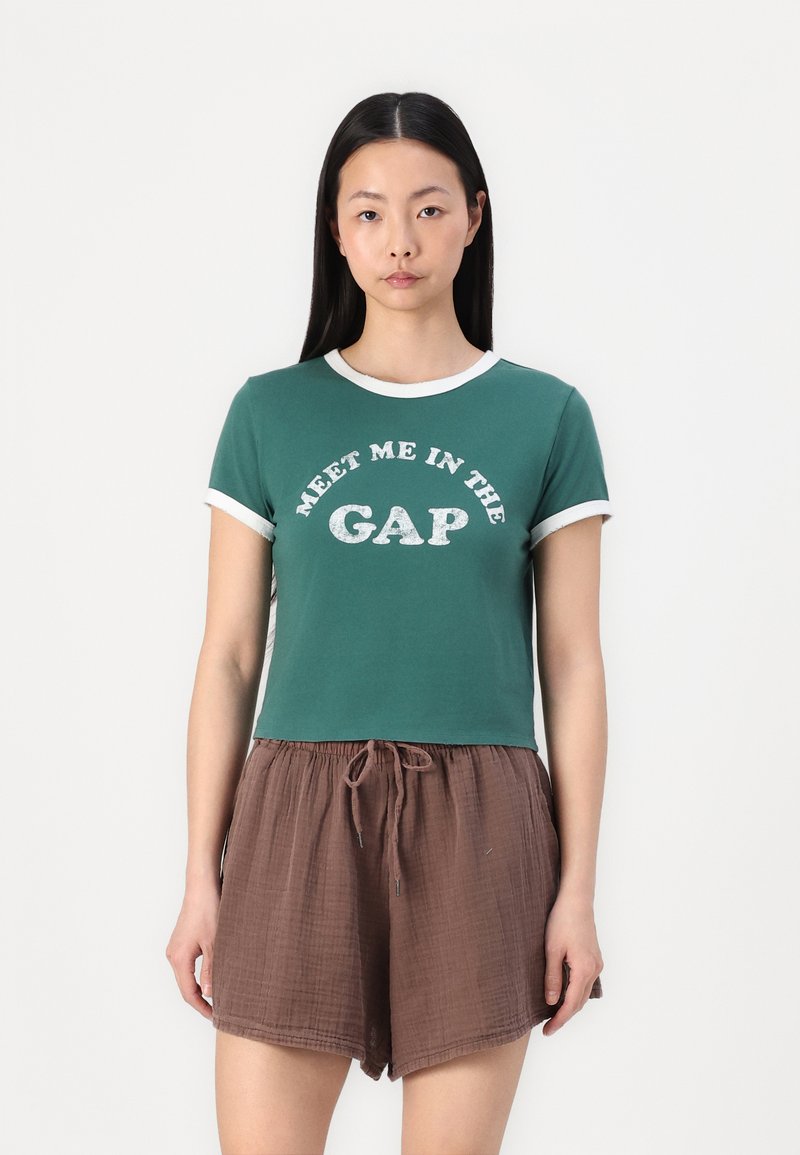 Camiseta verde de manga corta con acentos en blanco y el texto "ENCUÉNTRAME EN LA GRIETA", combinada con pantalones cortos marrones texturizados que tienen cordón.