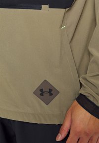 Khaki-takki mustilla yksityiskohdilla, jossa on timantin muotoinen logo-patch ja kontrastiväriset tikkaukset; sileä tekstuuri, joustavat hihansuut.