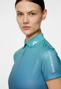 Polo shirt in een gradient teal stof, met een zachte kraag, knoopsluiting en witte branding op de schouder. Glad textuur.