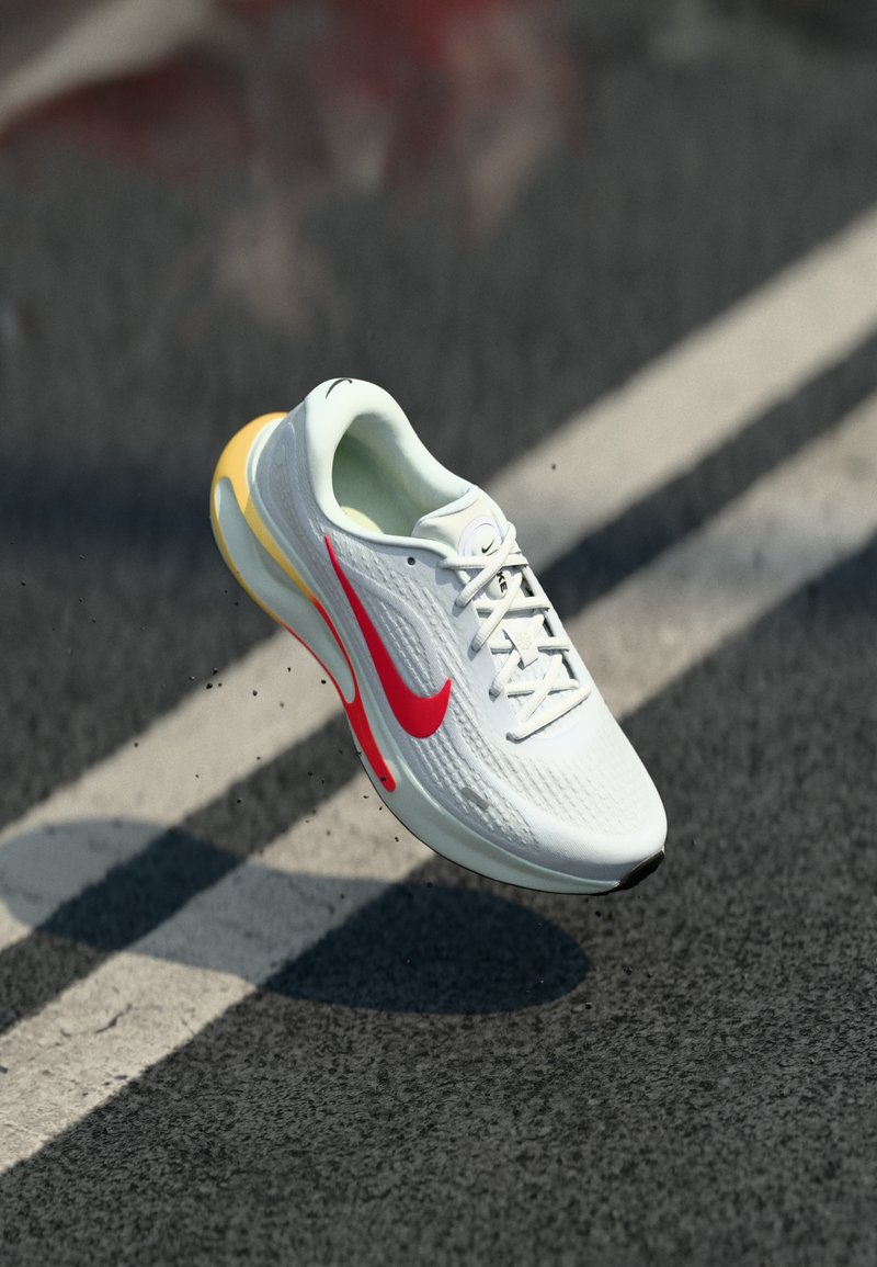 Chaussure de course Nike blanche et rouge suspendue au-dessus de l'asphalte avec des marquages de voie blancs, projetant une ombre au sol.