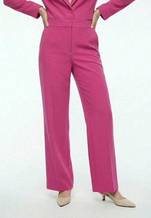 Femme portant un pantalon large taille haute rose vif avec une veste assortie et des escarpins beiges à bout pointu et talons bas.