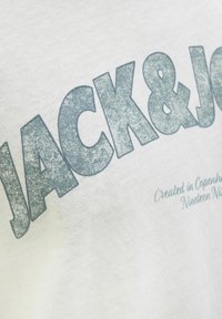 Balta kokvilnas T-krekls ar lieliem, mirdzošiem zila krāsas burtiem "JACK & JONES" un mazākiem burtiem apakšā, kas saka "Izveidots Kopenhāgenā 1990. gadā."
