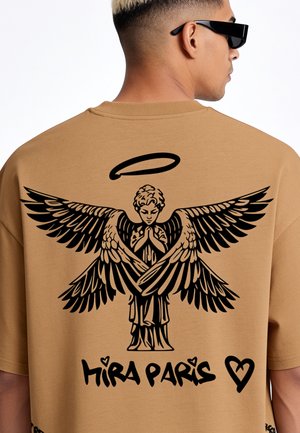 Mann mit kurzen Haaren und Sonnenbrille, der ein sandfarbenes Shirt mit schwarzer Grafik eines betenden Engels mit Flügeln, Heiligenschein, "Mira Paris" Text und Herzsymbol trägt.