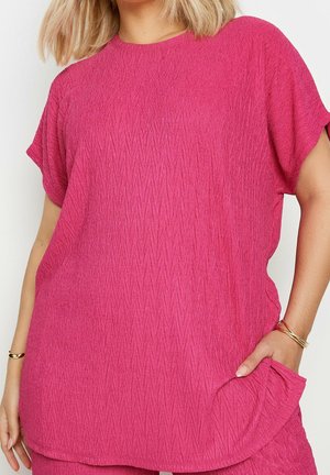 Kobieta ubrana w teksturowany, krótkim rękawem, fuksjowy top z okrągłym dekoltem, jedną ręką w kieszeni i złotymi bransoletkami na nadgarstkach.