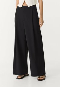 Zwarte wijde broek met een platte tailleband, gemaakt van gladde stof. Kenmerkt een strakke lijn en een losse pasvorm. Gecombineerd met platte sandalen met bandjes.