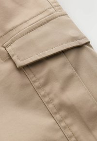 Beżowe spodnie typu cargo z tkaniny, zbliżenie pokazujące prostokątne kieszenie z klapką, widoczne podwójne przeszycia oraz fakturę tkaniny.