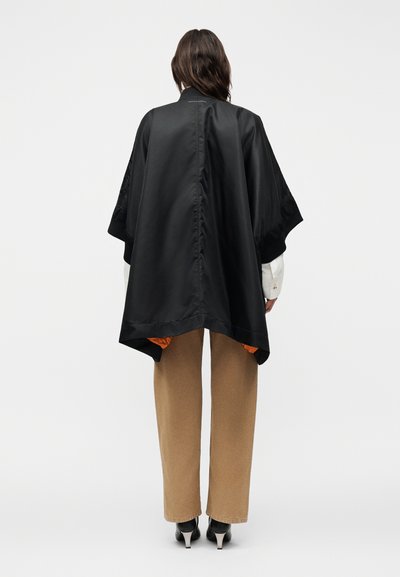 Άτομο που φοράει μια φαρδιά μαύρη κάπα με φαρδιά μανίκια πάνω από ένα λευκό πουκάμισο, μπεζ παντελόνια και μαύρα παπούτσια με τακούνι, στέκεται με την πλάτη γυρισμένη.