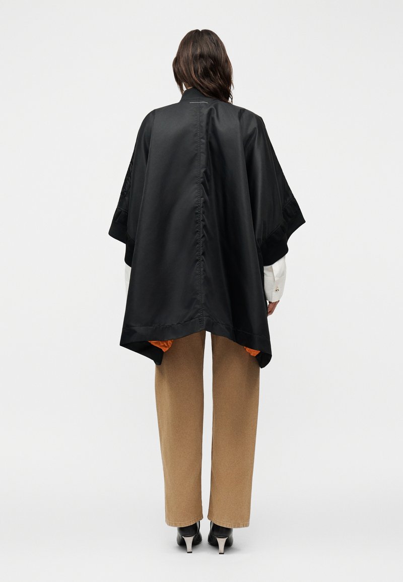 Personne portant une cape noire ample à larges manches par-dessus une chemise blanche, un pantalon beige et des chaussures noires à talons, debout de dos.