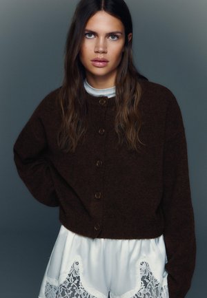 Brun oversized cardigan med rund halsudskæring og brune knapper, båret over hvide shorts med blonder, sat op mod en neutral baggrund.