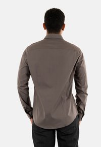 Hombre de espaldas que lleva una camisa gris de manga larga con botones y pantalones negros, frente a un fondo blanco liso.