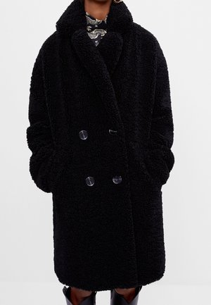 Femme portant un manteau teddy noir à double boutonnage avec de larges revers et les mains dans les poches, associé à un haut à motifs et des bottes noires.
