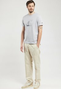 T-shirt en coton gris avec un graphique de voilier bleu, associé à un pantalon beige clair. L'ensemble est complété par des baskets blanches et beiges.