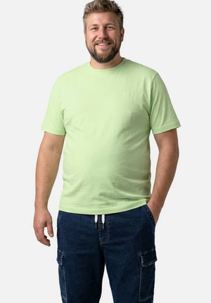 Usměvavý muž s vousy, oblečený v světle zeleném tričku s krátkým rukávem a tmavě modrých kalhotách s kapsami, stojící s jednou rukou v kapse.