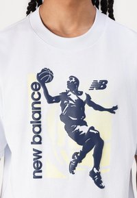 Balts kokvilnas t-krekls ar tumši zilu basketbolista siluetu, „new balance” tekstu un smalku dzeltenu fona rakstu.