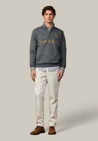 Grijze sweatshirt met gouden "HACKETT" logo, gestreepte kraag, gecombineerd met een blauw-wit gestreept shirt en crèmekleurige broeken. Bruine schoenen.