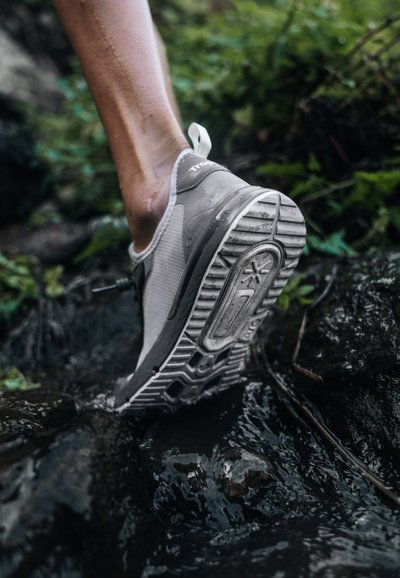 Gray athletic shoe with mesh upper and rugged rubber sole, submerged partially in water among rocks. Clear tread pattern visible.