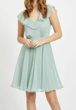 Kvinde iført lyseblå ærmeløs kjole med flæset halsudskæring og udvidet nederdel, stående med hænderne foldet foran.