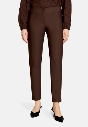 Femme portant un pantalon marron ajusté à jambes droites avec un bouton sur le devant et des escarpins noirs à bout pointu et talon bas sur fond blanc.