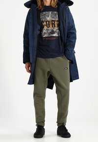 Marinblå lång rock över en marinblå t-shirt med trycket "CORE", olivgröna joggers och svarta sneakers med logotyp på joggers.