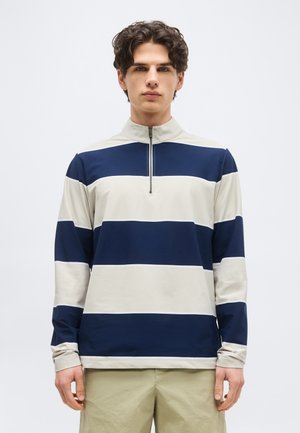 Jeune homme aux cheveux bouclés foncés portant un pull à larges rayures beige et bleu marine à manches longues avec une fermeture éclair à l'avant.