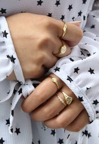 Drei goldene Ringe mit unterschiedlichen Designs, darunter ein Herz, ein florales Motiv und ein glatter Ring, die an Fingern vor einem weißen, sternmusternden Stoff getragen werden.