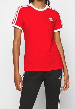 Osoba nosząca czerwoną koszulkę Adidas z białymi paskami i czarne spodnie dresowe Adidas z białymi paskami na białym tle.