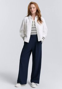 Balta džinsinė striukė dėvima ant juodai baltos dryžuotos palaidinės, derinant su tamsiai mėlynomis plačiomis kelnėmis ir baltais sportbačiais, stovint prie šviesaus fono.