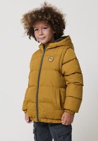 Chlapec s kučeravými vlasmi v hojdacej žltej puffa bunde s kapucňou a tmavých cargo nohaviciach, stojí na svetlom pozadí.