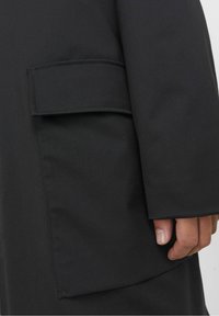 Black coat featuring a patch pocket, smooth fabric texture, and a loose fit. The sleeve is slightly flared at the wrist.