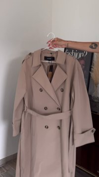 Bēšas krāsas divrindu tranča mētelis ar jostu, lieliem atlokiem un pogu akcentiem. Izgatavots no gludas auduma, ar strukturētām pleciem un piedurknēm.