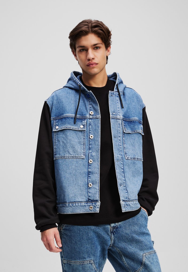 Denim jack met capuchon, lichtblauwe stof, met drukknopen aan de voorkant, twee borstzakken, gedragen over een zwart sweatshirt. Casual ontwerp.