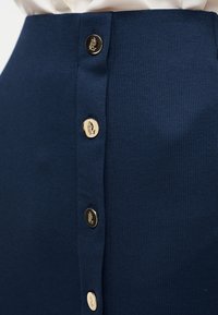 Φούστα σε ναυτικό μπλε με υφή και κάθετη σειρά από τέσσερα χρυσά κουμπιά χαραγμένα με λογότυπο, φορεμένη με λευκό ρούχο που έχει μπει μέσα από πάνω.