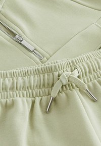 Lysgrønne sweatpants har et elastisk taljebånd med snøre og en lynlåslomme, lavet af blødt tekstureret stof.