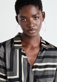 Camicia nera e panna con motivo a righe geometriche e colletto. Modella con orecchini a cerchio, sfondo neutro.