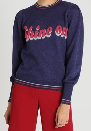 La personne porte un pull bleu marine avec le texte rouge et blanc « Shine On » et un pantalon rouge, montrant sa main droite ornée de bagues.