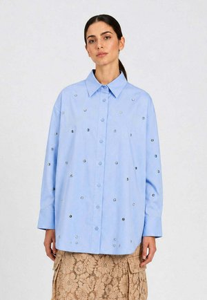 Donna che indossa una camicia oversize azzurro chiaro con borchie d'argento sparse e una gonna in pizzo floreale beige con tasche, in piedi davanti a uno sfondo semplice.