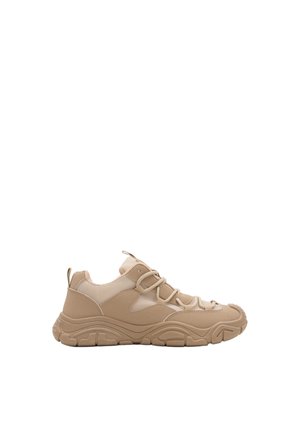 Zapatilla deportiva beige con suela gruesa y texturizada, parte superior de malla y ante, y cierre de cordones, vista desde el lado izquierdo sobre fondo blanco.