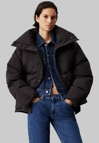 Zwarte pufferjas met een grote kraag, voorzien van een ritssluiting en knoopdetails, gedragen over een blauwe spijkerjack. Zacht en warm.