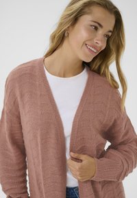 Femme souriante, portant un haut blanc sous un cardigan tricoté en rose poussiéreux texturé, avec de longs cheveux blonds et des boucles d'oreilles en or.