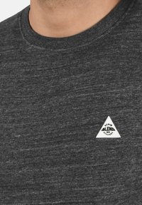 Crni melirani T-shirt s okruglim izrezom i malim bijelim trokutastim logotipom na lijevoj strani prsima, izrađen od teksturirane tkanine.
