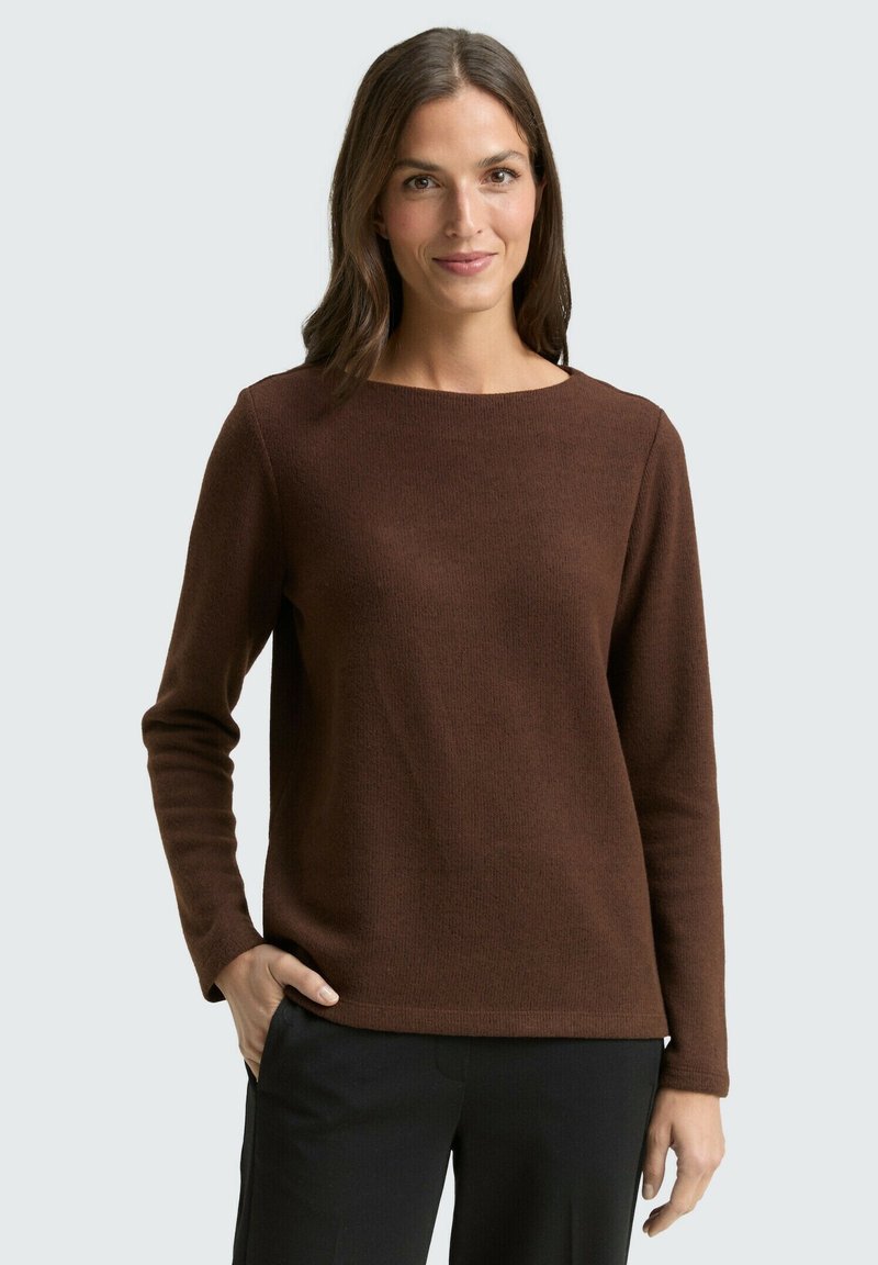 Femme aux cheveux bruns mi-longs portant un pull marron à manches longues et un pantalon noir, se tenant avec une main dans la poche.