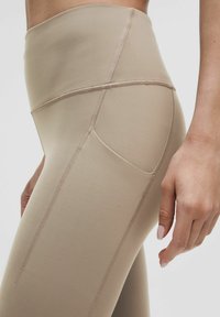 Leggings beige a vita alta realizzati in tessuto elasticizzato. Presentano cuciture piatte, tasca laterale e una texture liscia. Design aderente mostrato di lato.