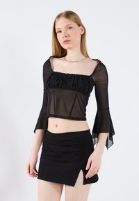 Čierny priesvitný cropped top s dlhými rukávmi a nazhromaždenými detailmi, doplnený čiernou mini sukňou so bočným rázporkom a hladkou textúrou.