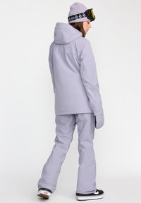 Tenue de ski violet clair composée d'une veste à capuche et d'un pantalon assorti. Le matériau semble lisse, avec un design minimaliste et des accents subtils.