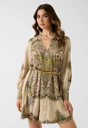 Jeune femme aux longs cheveux ondulés portant une robe ceinturée à manches longues avec des motifs cachemire et floraux verts et roses.