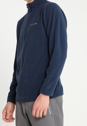 Person iført en navyblå Columbia trøje med lange ærmer og delvis lynlås samt grå bukser, stående mod en ensartet lys baggrund.