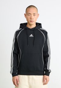 Homem a usar um hoodie preto da Adidas com riscas brancas nas mangas e logótipo no peito, de pé contra um fundo branco liso.