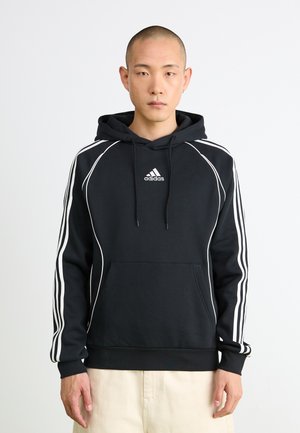 Bărbat îmbrăcat într-un hanorac negru Adidas cu dungi albe pe mâneci și logo pe piept, stând în fața unui fundal alb simplu.