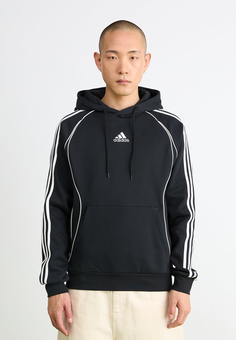 Homem a usar um hoodie preto da Adidas com riscas brancas nas mangas e logótipo no peito, de pé contra um fundo branco liso.