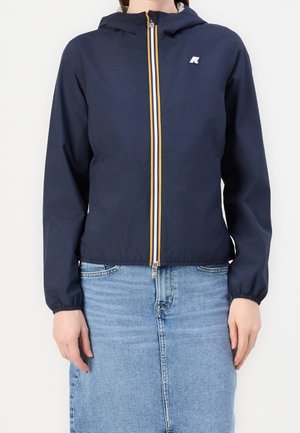 Vrouw draagt een marineblauwe capuchonvest met gele en witte ritssluitingsdetails, gecombineerd met een lichtblauwe denim rok.